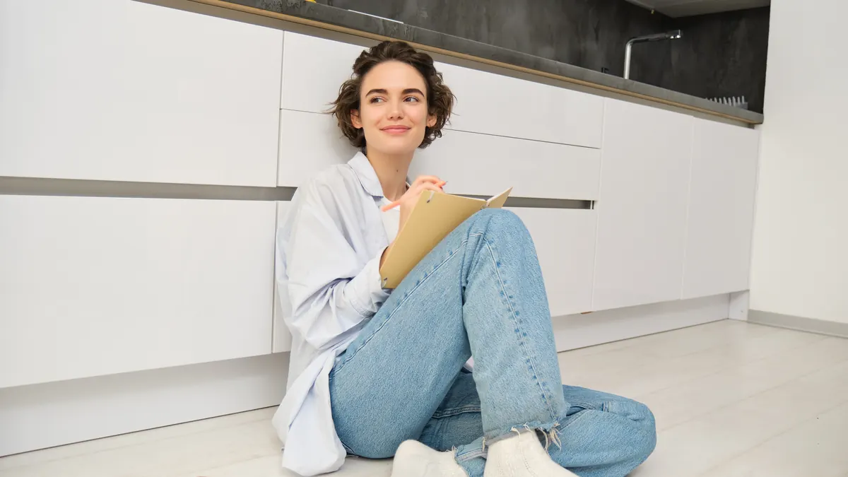A young woman sitting on a kitchen floor writing in her planner